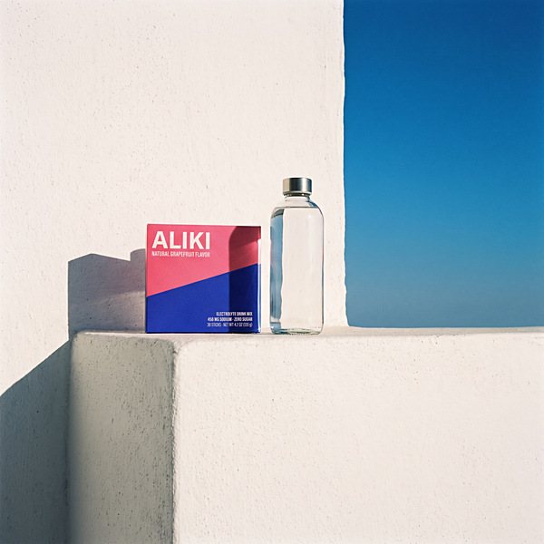 Closed ALIKI carton and a clear bottle of water on a whitewashed ledge at 9 am, long shadow