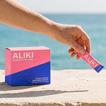 Hand lifting one ALIKI stick beside the closed carton on a sunlit ledge