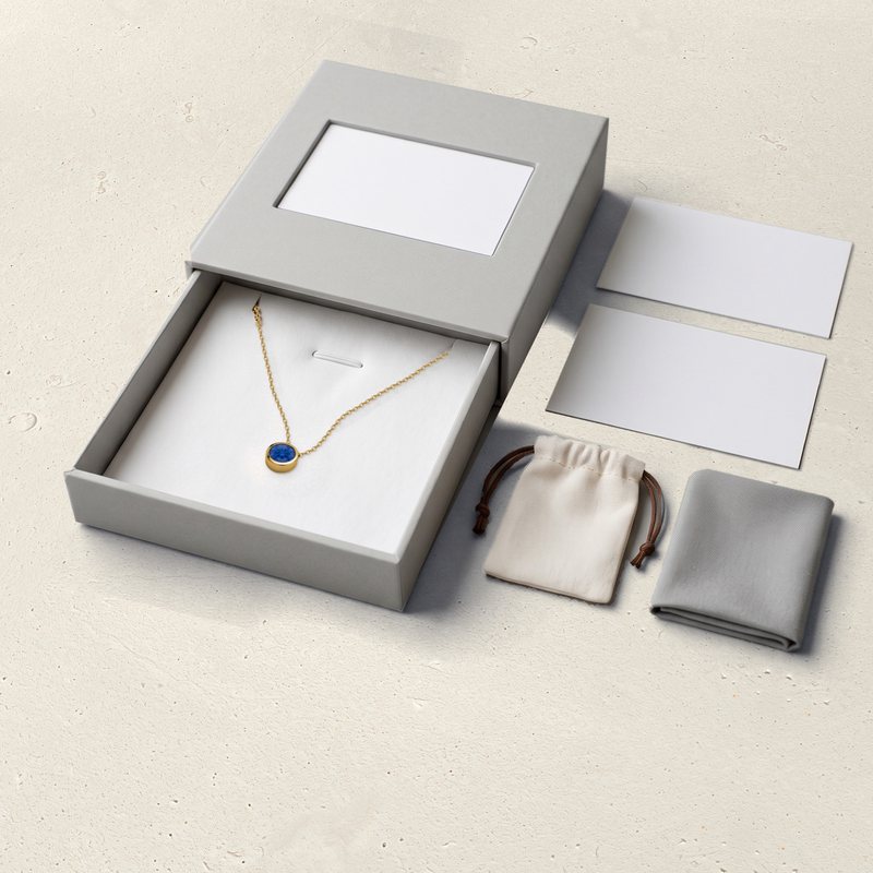 The Corund slide box open on pale limestone: the pendant on its insert, blank cards, a cotton pouch and a cloth